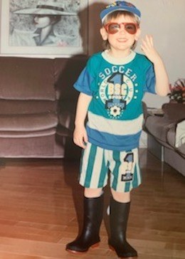 Un jeune Mathieu portant un chapeau, des lunettes de soleil, un t-shirt de football, un short rayé et des bottes de pluie